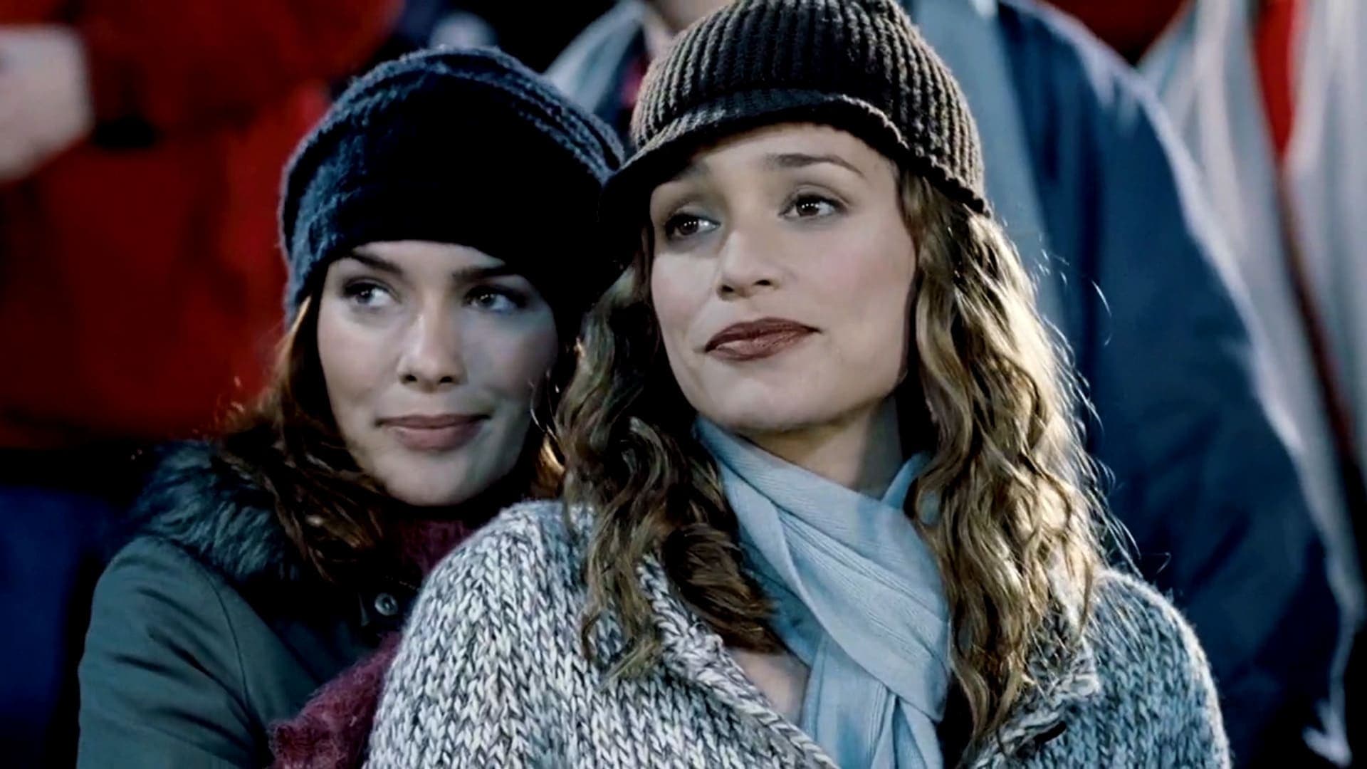 One woman seems to be hugging another, as they look into something. They both have warm clothes on. There's people behind them, from the movie one can know they're in a stadium.