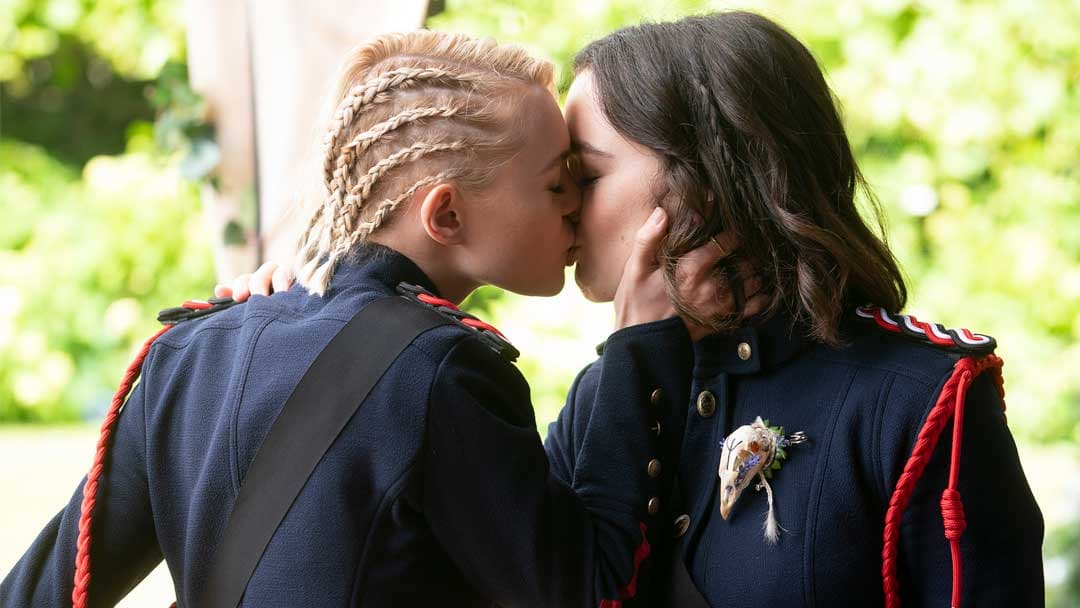 A blonde girl with braided hair and a brunette kiss. They're both wearing a dark blue uniform, which looks military.