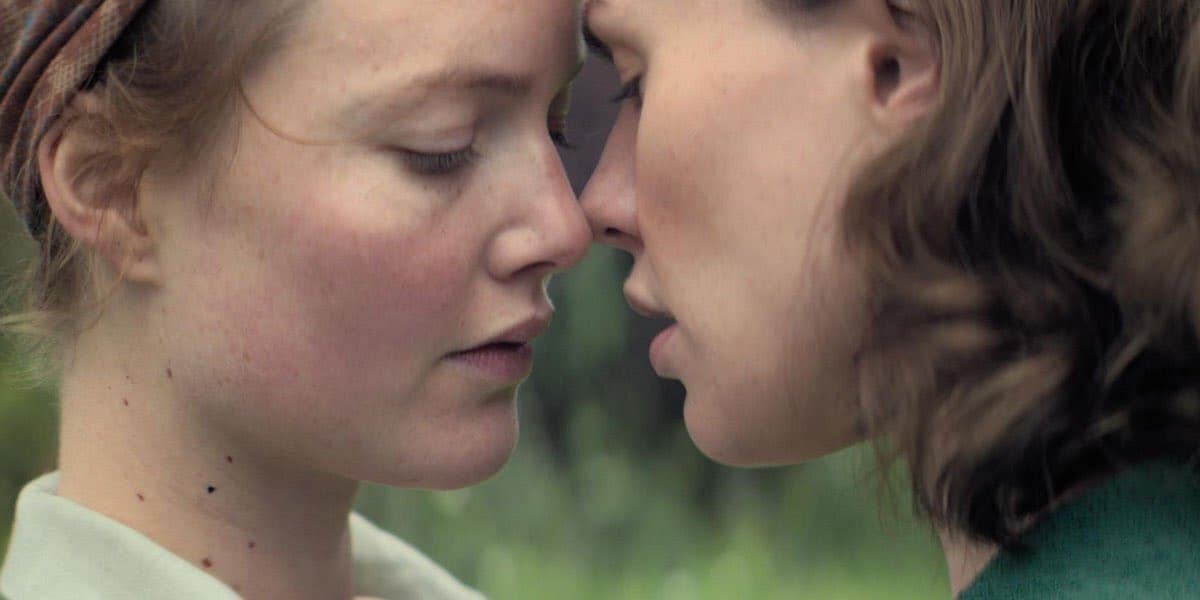 Two woman are very close, face to face, their foreheads almost touching. They seem about to kiss.