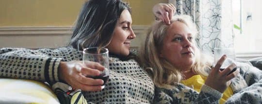 Gunhild, blonde, and Luna, brunette, are sitting on a couch, embracing each other. Luna is playing with Gunhild's hair. They both have a glass of wine and are dressed for cold climate.